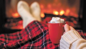 Woman with cup of hot cocoa and marshmallow warming legs in winter white socks near fireplace flame, covered christmas plaid